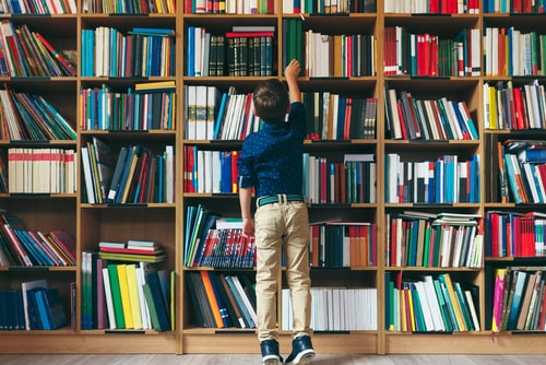 Boy stretches after a book on multi colored bookshelf in library.